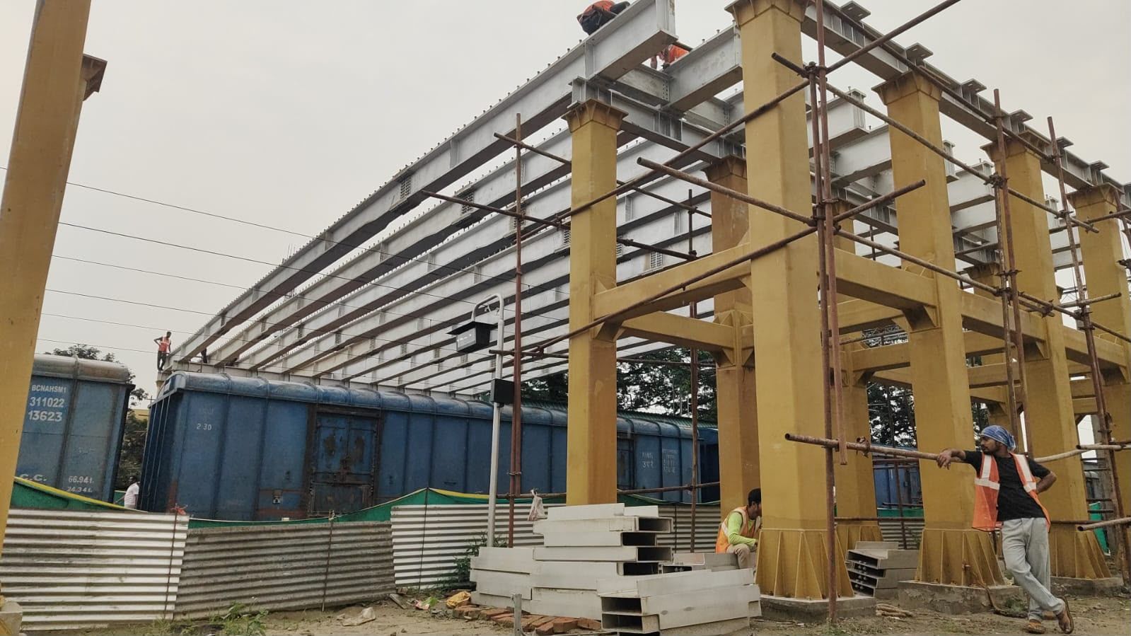 Structural steel frame with yellow-painted columns and grey roof girders, beside railway wagons