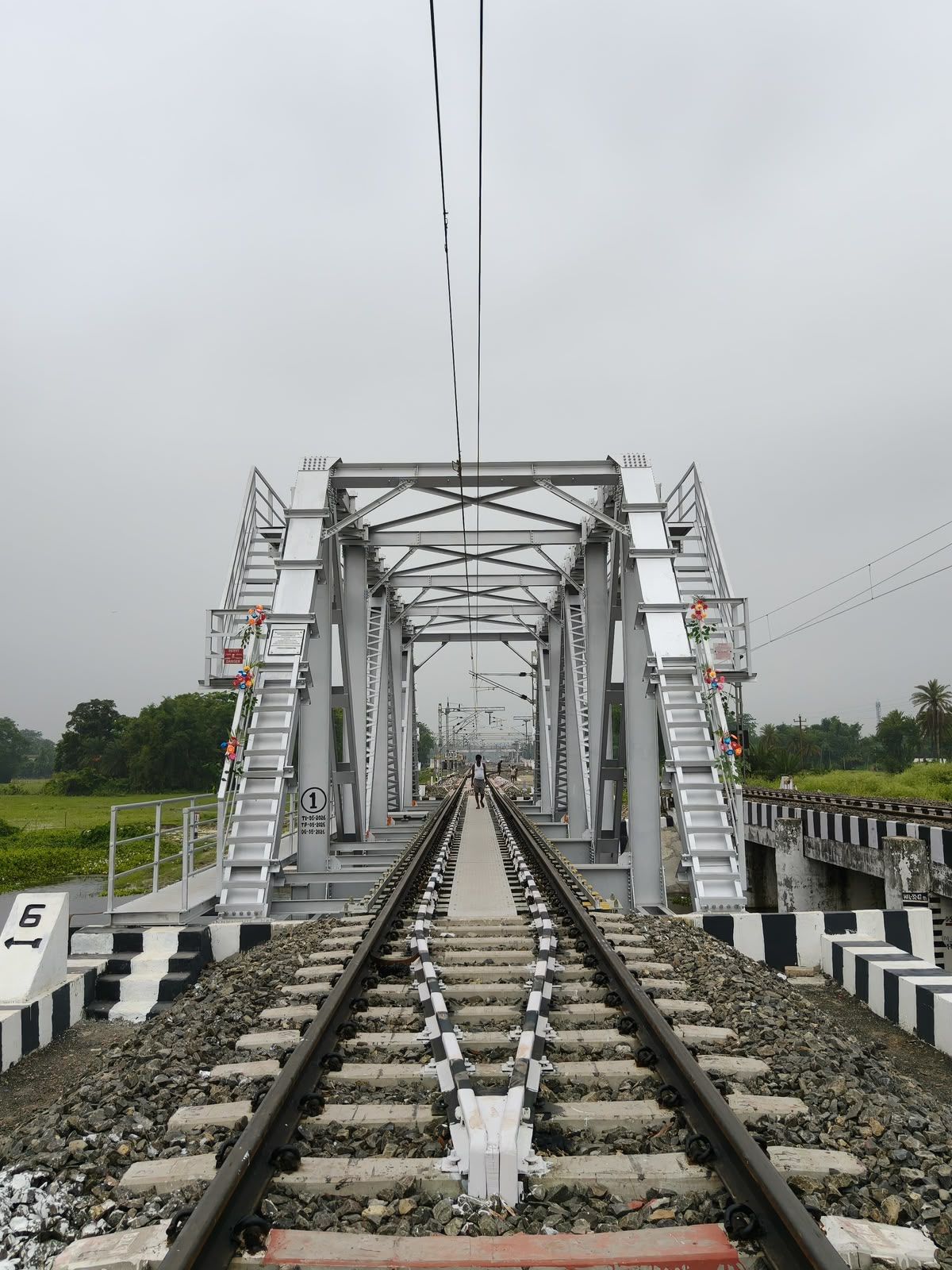 View along a railway track through a painted steel truss bridge, under overhead electric lines
